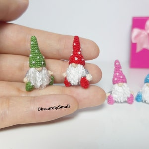 May include: Four miniature crocheted gnomes with different coloured hats. The gnomes are sitting on a hand. The gnomes are wearing white beards and have red, green, pink, and blue hats.