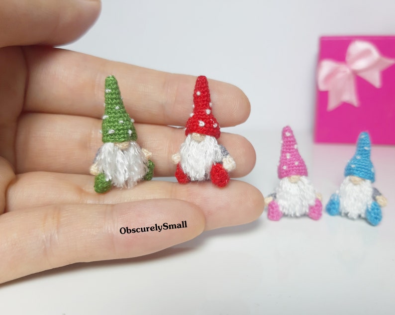 May include: Four miniature crocheted gnomes with different coloured hats. The gnomes are sitting on a hand. The gnomes are wearing white beards and have red, green, pink, and blue hats.