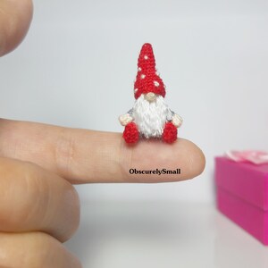 May include: A tiny crocheted gnome with a red and white dotted hat, wearing a blue and white striped shirt and red trousers. The gnome is sitting on a finger.