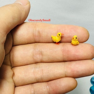 May include: Two tiny crocheted yellow ducklings with orange beaks and feet. The ducklings are sitting on a person's hand.