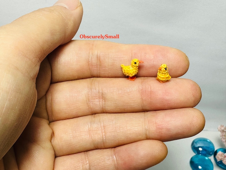 May include: Two tiny crocheted yellow ducklings with orange beaks and feet. The ducklings are sitting on a person's hand.