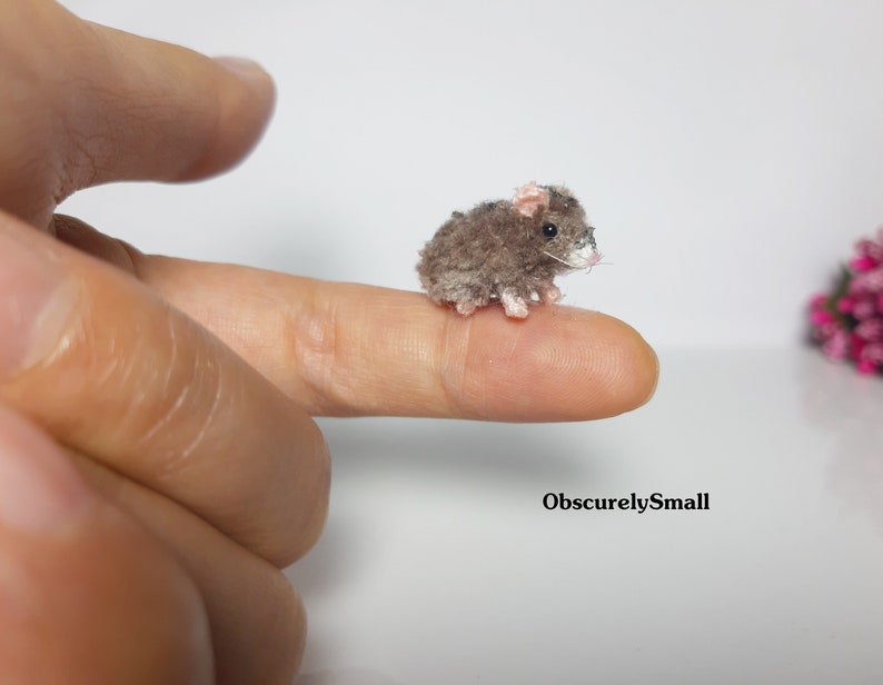 May include: A small, gray crocheted mouse with pink ears. The mouse is sitting on a finger.