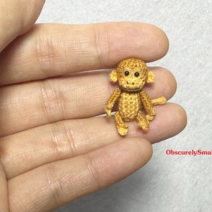 May include: A small crocheted monkey toy with brown fur, black eyes, and a light brown nose. The monkey is sitting on a hand.