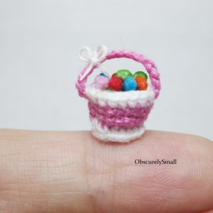 Pu&ograve; includere: Un cestino in miniatura di crochet rosa e bianco pieno di palline colorate in crochet. Il cestino &egrave; appoggiato su un dito.