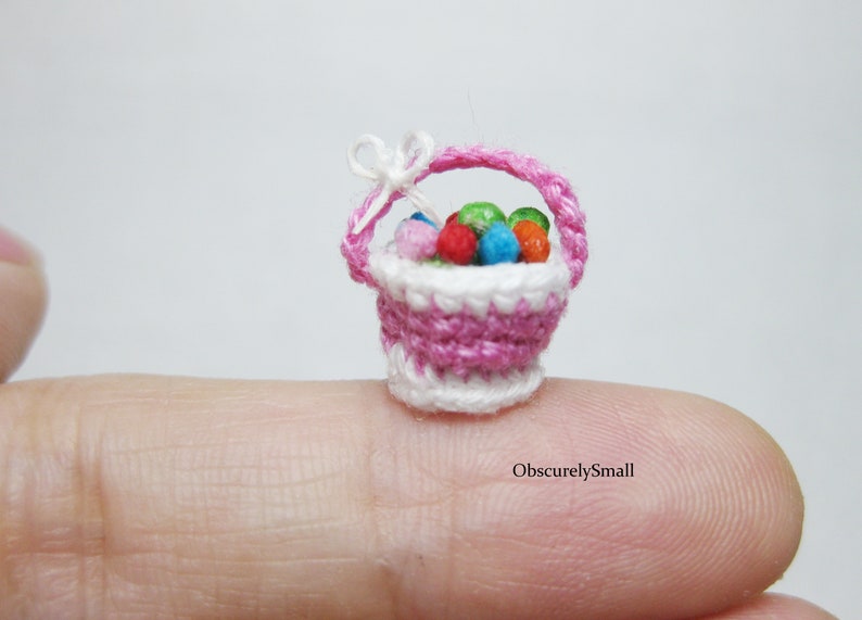 Pu&ograve; includere: Un cestino in miniatura di crochet rosa e bianco pieno di palline colorate in crochet. Il cestino &egrave; appoggiato su un dito.