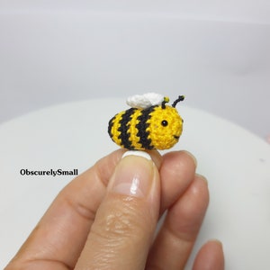 Op de afbeelding: Een kleine gehaakte bij met gele en zwarte strepen, witte vleugels en zwarte ogen. De bij wordt vastgehouden door een hand.
