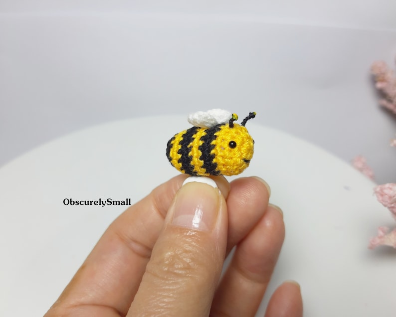 Op de afbeelding: Een kleine gehaakte bij met gele en zwarte strepen, witte vleugels en zwarte ogen. De bij wordt vastgehouden door een hand.