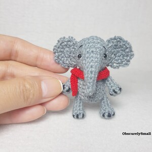 May include: A small, grey crocheted elephant figurine featuring large ears, a trunk, and a bright red scarf wrapped around its neck. The elephant has black bead eyes and small black stitched details on its feet. The text ObscurelySmall is visible.
