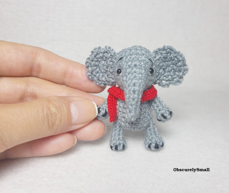 May include: A small, grey crocheted elephant figurine featuring large ears, a trunk, and a bright red scarf wrapped around its neck. The elephant has black bead eyes and small black stitched details on its feet. The text ObscurelySmall is visible.