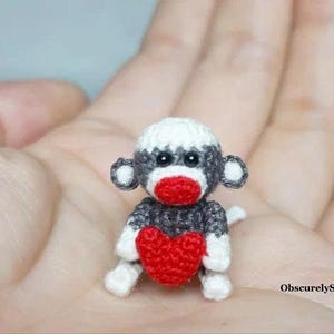 May include: A miniature crocheted monkey figurine featuring dark grey yarn with white accents on the head, ears, and feet, a bright red mouth area, and a red heart held in its arms. The text ObscurelySmall is printed in the lower right corner.