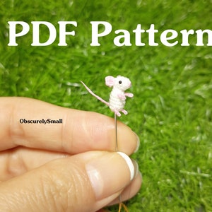 May include: PDF pattern for crocheting a tiny white mouse with pink ears. The mouse is sitting on a pin and is being held by a hand. The background is green grass.
