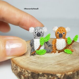Può includere: Due piccoli koala all'uncinetto seduti su una fetta di legno. Il koala a sinistra è grigio e il koala a destra è marrone. Entrambi i koala tengono foglie verdi.