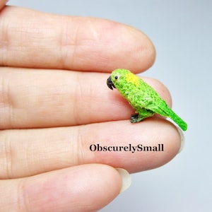 Op de afbeelding: Een kleine gehaakte papegaai met groene veren en een gele kop. De papegaai zit op een vinger.