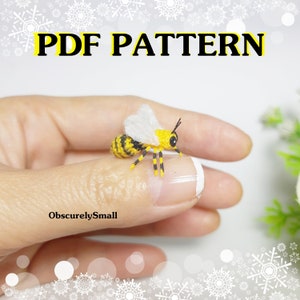 May include: A close-up of a crocheted bee with yellow and black stripes, white wings, and black bead eyes. The bee is sitting on a finger.