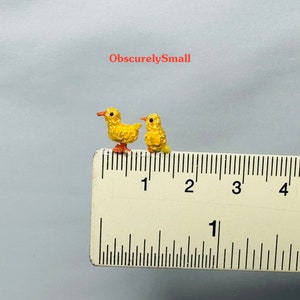 May include: Two small yellow crocheted chicks, each about 1 cm tall, are positioned on a ruler. The chicks are facing the right side of the image. The ruler is marked in inches and shows the chicks are less than 1 inch tall.