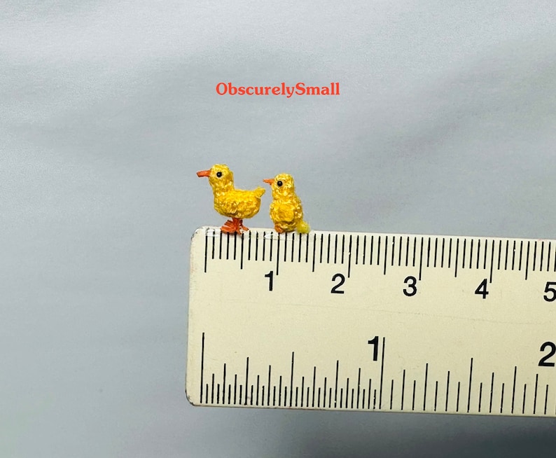 May include: Two small yellow crocheted chicks, each about 1 cm tall, are positioned on a ruler. The chicks are facing the right side of the image. The ruler is marked in inches and shows the chicks are less than 1 inch tall.