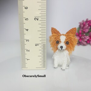 May include: A small crocheted dog toy, approximately 2 inches tall, with a white body and orange and brown fur. The dog is sitting with its paws tucked underneath it. A ruler is in the background for size reference.