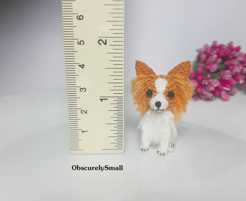 May include: A small crocheted dog toy, approximately 2 inches tall, with a white body and orange and brown fur. The dog is sitting with its paws tucked underneath it. A ruler is in the background for size reference.