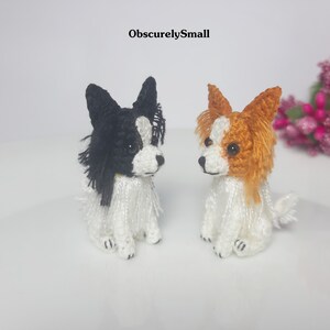 May include: Two small crocheted Papillon dogs, one black and white, the other brown and white, sitting on a white surface.  The text "ObscurelySmall" is visible above the dogs.