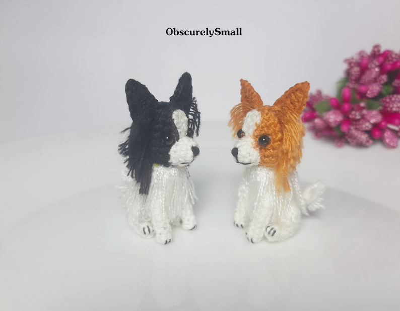 May include: Two small crocheted Papillon dogs, one black and white, the other brown and white, sitting on a white surface.  The text "ObscurelySmall" is visible above the dogs.