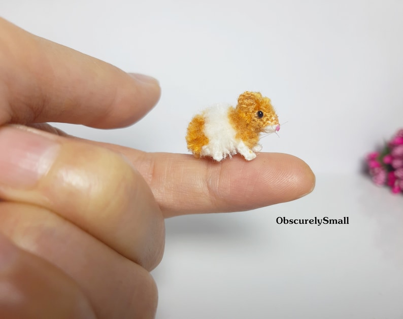 May include: A tiny crocheted hamster with brown and white fur, sitting on a finger. The hamster has a pink nose and small black eyes.