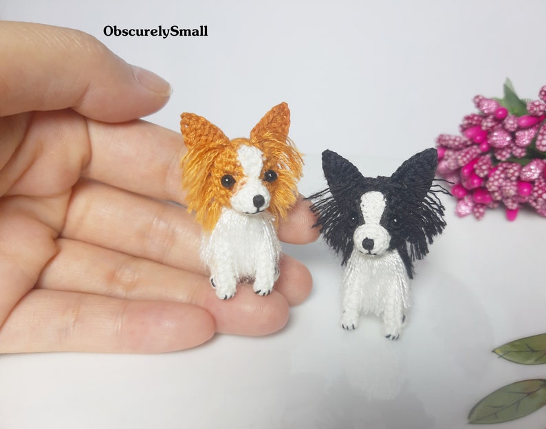 May include: Two small crocheted Papillon dogs, one with orange and white fur and the other with black and white fur. Both dogs are sitting with their ears perked up and looking at the camera.
