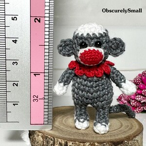 Puede incluir: Un peque&ntilde;o mono de crochet gris con detalles blancos y un collar de crochet rojo. El mono mide aproximadamente 5 cm de alto y est&aacute; de pie sobre una superficie de madera.