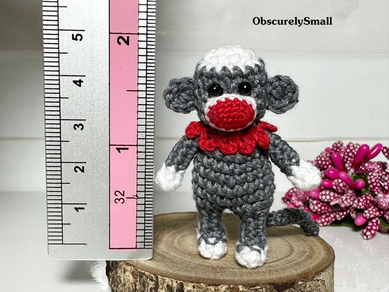 Puede incluir: Un peque&ntilde;o mono de crochet gris con detalles blancos y un collar de crochet rojo. El mono mide aproximadamente 5 cm de alto y est&aacute; de pie sobre una superficie de madera.