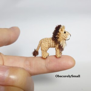 Pu&ograve; includere: Un piccolo leone all'uncinetto con una criniera marrone, in piedi su un dito. Il leone &egrave; color beige e marrone e ha un viso bianco.