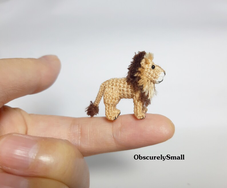 Pu&ograve; includere: Un piccolo leone all'uncinetto con una criniera marrone, in piedi su un dito. Il leone &egrave; color beige e marrone e ha un viso bianco.