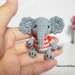 May include: A small, grey crocheted elephant figurine wearing a red and white patterned shirt. The elephant has large ears, a trunk, and black bead eyes. The text "ObscurelySmall" is printed in the bottom right corner of the image.