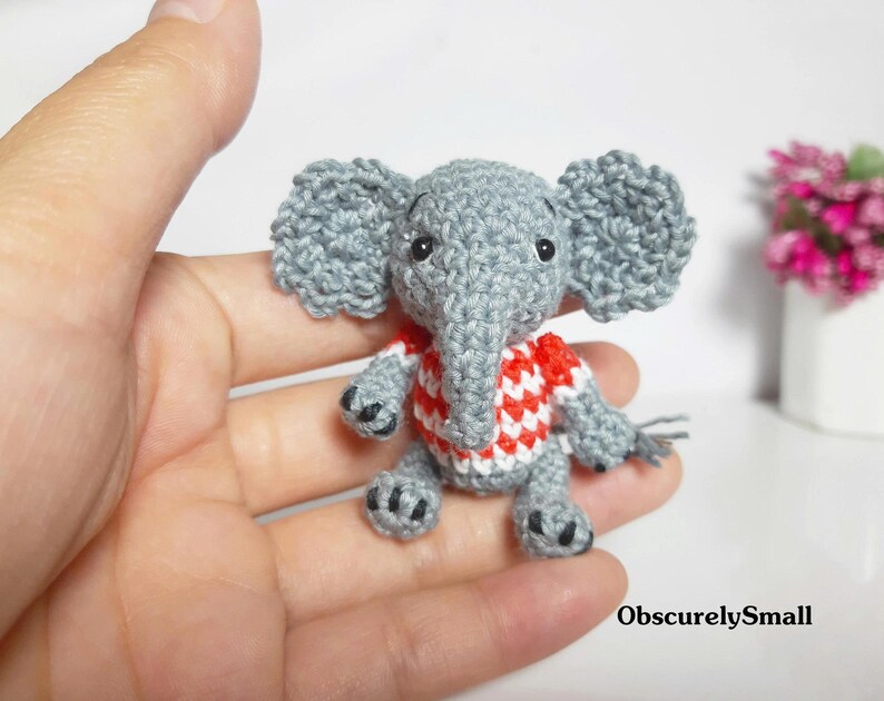 May include: A small, grey crocheted elephant figurine wearing a red and white patterned shirt. The elephant has large ears, a trunk, and black bead eyes. The text "ObscurelySmall" is printed in the bottom right corner of the image.