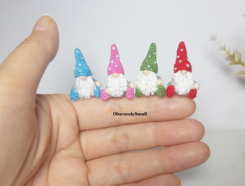 May include: Five miniature crocheted gnomes in different colours, including blue, pink, green, and red. Each gnome has a white beard and a pointed hat with white polka dots.