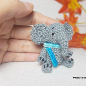May include: A small gray crocheted elephant figurine featuring large ears, a trunk, and a bright blue scarf around its neck. The elephant has black embroidered eyes and black stitched details on its feet. The text ObscurelySmall is visible.