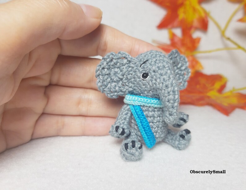 May include: A small gray crocheted elephant figurine featuring large ears, a trunk, and a bright blue scarf around its neck. The elephant has black embroidered eyes and black stitched details on its feet. The text ObscurelySmall is visible.
