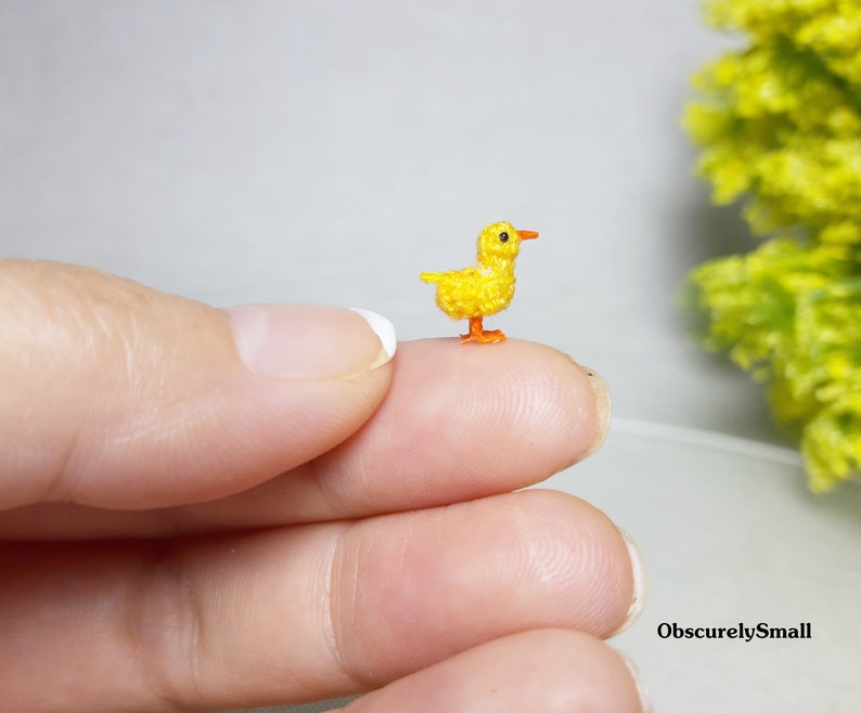 May include: A tiny crocheted yellow chick with orange feet and beak. The chick is sitting on a finger.