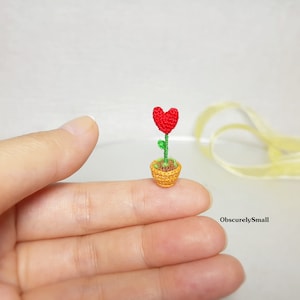 May include: A tiny crocheted heart-shaped flower in a yellow pot, held in a person's hand. The flower is red and the stem is green.