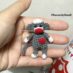 Puede incluir: Un peque&ntilde;o mono de crochet gris con la nariz roja y un coraz&oacute;n rojo en el pecho. El mono sostiene un coraz&oacute;n rojo en sus manos. El mono est&aacute; sentado en una mano.