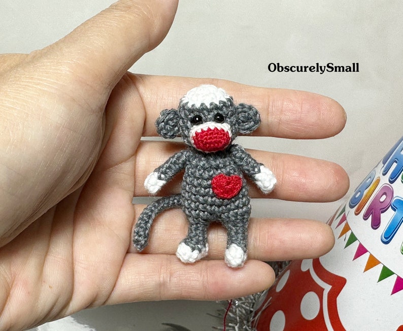 Puede incluir: Un peque&ntilde;o mono de crochet gris con la nariz roja y un coraz&oacute;n rojo en el pecho. El mono sostiene un coraz&oacute;n rojo en sus manos. El mono est&aacute; sentado en una mano.