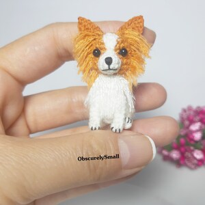 May include: A small crocheted dog with white fur and orange ears. The dog is standing on a hand.