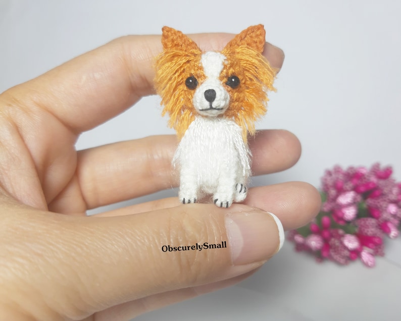 May include: A small crocheted dog with white fur and orange ears. The dog is standing on a hand.
