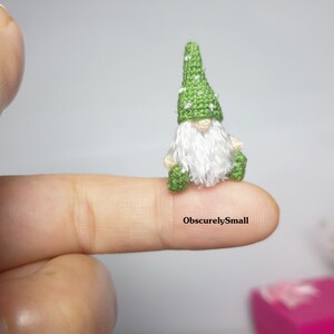 May include: A tiny green gnome with a white beard and a green hat with white dots sits on a finger. The gnome is very small, and the text "ObscurelySmall" is visible on the finger.