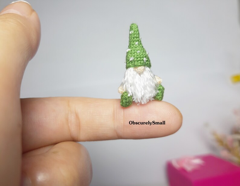 May include: A tiny green gnome with a white beard and a green hat with white dots sits on a finger. The gnome is very small, and the text "ObscurelySmall" is visible on the finger.