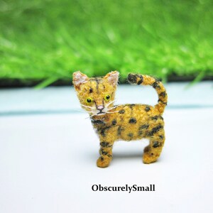 Pu&ograve; includere: Una piccola figurina di un gatto tigrato marrone e nero all'uncinetto con occhi gialli. Il gatto &egrave; in piedi su una superficie bianca.