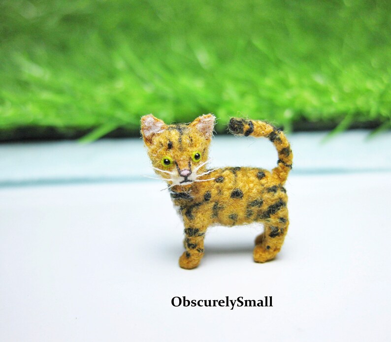 Pu&ograve; includere: Una piccola figurina di un gatto tigrato marrone e nero all'uncinetto con occhi gialli. Il gatto &egrave; in piedi su una superficie bianca.