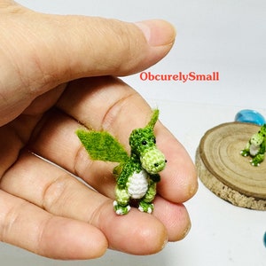 May include: Two small green crocheted dragons with white accents. The dragons are sitting on a wooden surface and a hand is holding one of the dragons.