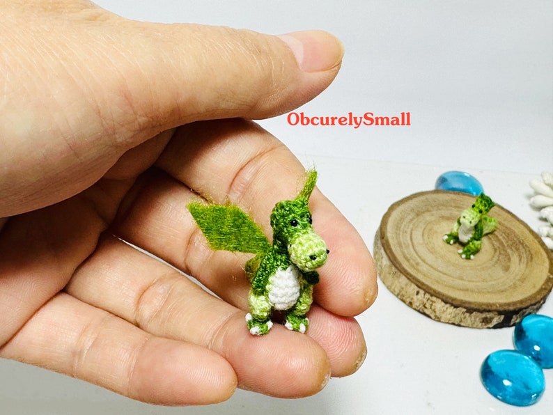 May include: Two small green crocheted dragons with white accents. The dragons are sitting on a wooden surface and a hand is holding one of the dragons.