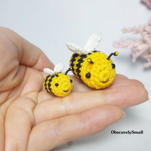 Op de afbeelding: Twee gehaakte hommels, een groter dan de andere, zitten in een hand. De grotere hommel is geel met zwarte strepen en witte vleugels. De kleinere hommel is geel met zwarte strepen en witte vleugels. Beide hommels hebben zwarte ogen en een glimlachende mond.