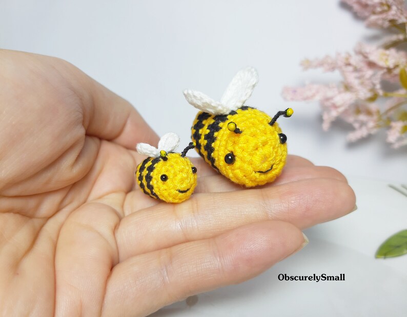 Op de afbeelding: Twee gehaakte hommels, een groter dan de andere, zitten in een hand. De grotere hommel is geel met zwarte strepen en witte vleugels. De kleinere hommel is geel met zwarte strepen en witte vleugels. Beide hommels hebben zwarte ogen en een glimlachende mond.