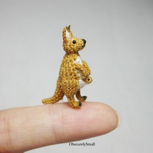 May include: A small crocheted kangaroo with a joey in its pouch. The kangaroo is brown and white, and the joey is white. The kangaroo is standing on a finger.